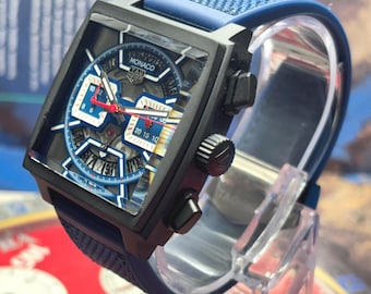 Puede incluir: Un reloj TAG Heuer Monaco de caja cuadrada con marco negro y esfera esqueletizada con detalles en azul y blanco. El reloj tiene una correa de caucho azul texturizada, manecillas rojas y dos subesferas. La palabra MONACO aparece en la esfera.