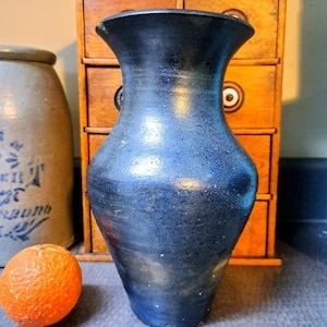 May include: A dark blue ceramic vase with a flared rim and a textured surface. The vase is set against a wooden cabinet and a stoneware jar. An orange fruit sits in front of the vase.