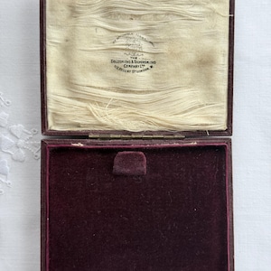 May include: A rectangular hinged jewelry box with a dark burgundy exterior. The interior lid features cream-colored silk lining with the printed text: The Goldsmiths & Silversmiths Company Ltd, 112 Regent St London, W. The base has dark purple velvet.