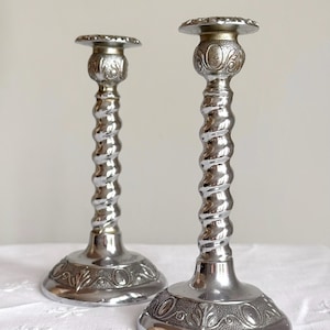 Op de afbeelding: Twee zilverkleurige metalen kandelaars met een gedraaid spiraalvormig ontwerp. Elke kandelaar heeft een ronde voet met reliëf van bloem- en ovaalmotieven, een textuur op de hals en een platte, ronde bovenplaat voor een kaars.