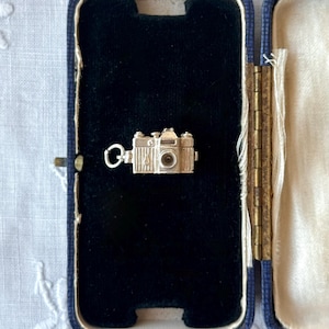 Op de afbeelding: Een miniatuur zilverkleurige camera-bedel met een gedetailleerde lens, zoeker en een kleine ronde ring aan de zijkant, gepresenteerd in een rechthoekig marineblauw scharnierend doosje met een zwarte fluwelen voering aan de binnenzijde.