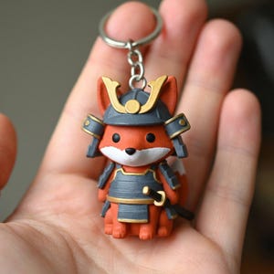 May include: A keychain featuring a small, orange fox figurine dressed in dark gray and gold samurai armor. The fox has a white muzzle, black eyes, and a small sword at its side. The figure is attached to a silver-colored metal split ring.