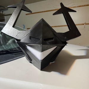 Puede incluir: Un casco negro angular con dos grandes cuernos que apuntan hacia arriba con puntas en forma de flecha. El casco tiene una sección frontal puntiaguda con ranuras oculares horizontales y una abertura bucal blanca, ancha y triangular.