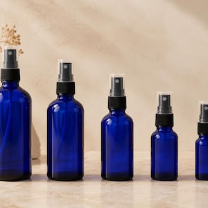 Peut inclure: Cinq flacons pulvérisateurs en verre bleu cobalt de différentes tailles, disposés en rangée. Chaque flacon est doté d'un pulvérisateur noir avec un couvercle en plastique transparent. Les flacons sont sur une surface claire avec un fond neutre.