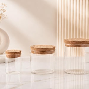 Peut inclure: Trois bocaux de rangement cylindriques en verre transparent de hauteurs différentes, chacun surmonté d'un couvercle en liège rond de couleur naturelle. Les bocaux sont vides et présentent des parois lisses et transparentes, alignés.
