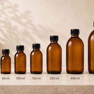 Op de afbeelding: Een rij van zeven amberkleurige glazen flessen met zwarte schroefdoppen, gerangschikt op grootte van links naar rechts. De flessen zijn gelabeld met inhoudsmaten van 30 ml, 60 ml, 100 ml, 125 ml, 250 ml, 500 ml en 1000 ml, wat een duidelijke groei toont.