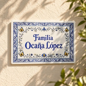 Könnte beinhalten: Ein rechteckiges Keramik-Hausschild mit weißem Hintergrund und einem blauen Zierrahmen, der florale Muster und gelbe Schmetterlinge zeigt. Der Text "Familia Ocaña López" ist mittig in einer blauen Serifenschrift aufgedruckt.