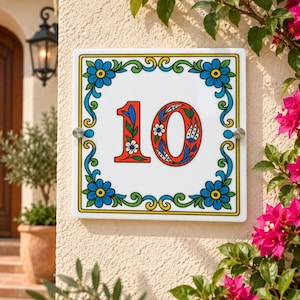 May include: A square ceramic house number plaque displaying the number 10 in a bold red font with intricate floral patterns inside the digits. The border features blue flowers and green vines on a white background, secured by two silver metal mounts.