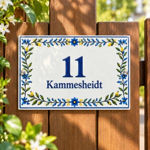 Könnte beinhalten: Ein rechteckiges weißes Keramik-Hausschild mit der Nummer 11 und dem Schriftzug Kammesheidt in dunkelblauer Farbe. Das Schild ist mit einem handgemalten Rand aus blauen Blumen, grünen Blättern und gelben Akzenten verziert und mit Schrauben befestigt.