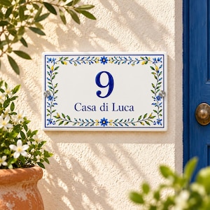 Könnte beinhalten: Ein rechteckiges Hausnummernschild aus weißer Keramik mit der blauen Ziffer 9 und dem Schriftzug Casa di Luca in blauer Farbe. Das Schild ist mit einem bemalten Rand aus blauen Blumen, grünen Blättern und gelben Akzenten verziert und mit silbernen Metallschrauben befestigt.