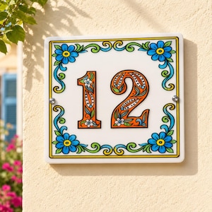 Peut inclure: Une plaque de numéro de maison carrée en céramique affichant le nombre 12 en orange avec des motifs floraux complexes. La bordure présente des fleurs bleues et des vignes vertes sur fond blanc, fixée par deux vis métalliques argentées.