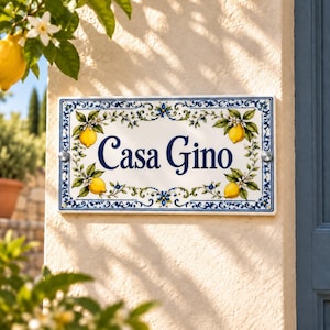 Puede incluir: Un letrero rectangular de cerámica para casa con el texto Casa Gino en letra cursiva azul oscuro. El borde está decorado con limones amarillos, hojas verdes, flores blancas y motivos florales azules sobre un fondo blanco.