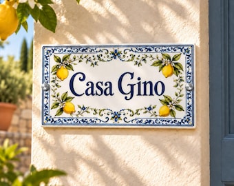 Peut inclure: Une plaque de maison rectangulaire en céramique portant l'inscription Casa Gino en lettres cursives bleu foncé. La bordure est ornée de citrons jaunes, de feuilles vertes, de fleurs blanches et de motifs floraux bleus sur fond blanc.