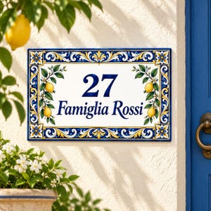 Könnte beinhalten: Ein rechteckiges Keramik-Hausschild mit der Nummer 27 und dem Text Famiglia Rossi in dunkelblauer Schrift. Der Rand ist mit gelben Zitronen, grünen Blättern und weißen Blüten auf weißem Grund sowie blau-gelben Verzierungen geschmückt.