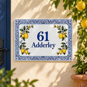 Puede incluir: Un letrero rectangular para casa con fondo blanco, que muestra el número 61 y el texto Adderley en azul. El letrero está decorado con un borde floral azul e ilustraciones de limones amarillos con hojas verdes a los lados.