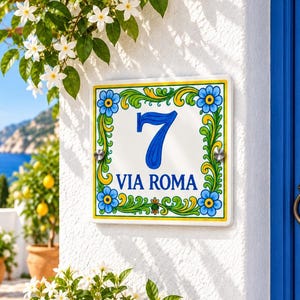 Peut inclure: Une plaque de numéro de maison carrée en céramique affichant le chiffre 7 et le texte VIA ROMA en bleu. Le motif présente une bordure décorative de fleurs bleues et de feuilles vertes sur fond blanc, fixée par deux boulons en métal argenté.