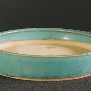 May include: A shallow, round ceramic bonsai pot with a vibrant turquoise blue glaze on the exterior and a smooth, light beige interior. The pot features a flat rim and two drainage holes visible at the bottom of the basin, showcasing a clean design.