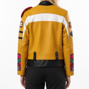 May include: A mustard yellow jacket with a black collar and hem, featuring a horizontal white stripe across the back. The sleeves display colorful graphic patterns, patches, and black stripes. Two black tabs with buttons are positioned at the waist.