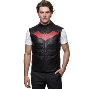 May include: A black leather vest featuring a high mock neck collar and a prominent red bat-shaped emblem on the chest. The vest has molded, segmented paneling across the torso, creating a structured appearance over a plain white short-sleeved t-shirt.