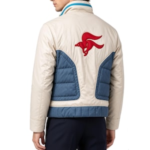 Peut inclure: Une veste beige avec des panneaux matelassés bleus sur le bas du dos et les côtés. La veste présente un col montant avec un liseré bleu et un grand logo renard rouge brodé dans le haut du dos. Le modèle inclut des manches longues.