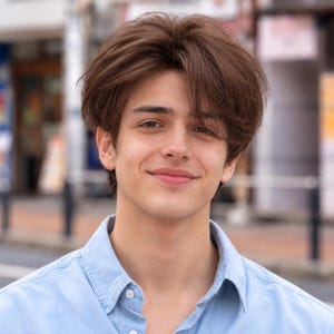 May include: A person with medium-length, layered brown hair styled with volume and a soft side-swept fringe. The individual is wearing a light blue collared button-down shirt with a visible white button at the neckline and a classic collar.