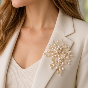 May include: A decorative brooch featuring a cluster of small, white, oval-shaped pearls attached to thin, gold-toned wire branches. The brooch is pinned to the lapel of a cream-colored blazer, showcasing a spray-like arrangement of pearls.