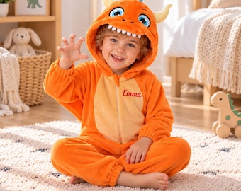 May include: A bright orange plush onesie featuring a hood with a monster face, white teeth, and two horns. The garment has a light beige panel on the chest with the name Emma embroidered in red script and a front zipper closure.