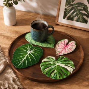 May include: A wooden tray displays a mug of coffee and four leaf-shaped coasters. The coasters are green, white, and pink, with detailed veining. A framed botanical print and a small vase are in the background.