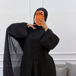 May include: A black abaya featuring a central vertical panel with matching fabric-covered buttons and wide sleeves. The garment is paired with a coordinating black hijab adorned with a white polka dot pattern, draped over the shoulders.