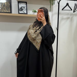 May include: A solid black abaya with long sleeves and a loose, flowing silhouette, paired with a square headscarf featuring a detailed brown, beige, and black paisley and geometric pattern that drapes over the shoulders and covers the head.