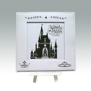 Pu&ograve; includere: Un biglietto bianco con una silhouette nera di un castello e il testo "Happily Ever After" in scrittura nera. Il biglietto &egrave; su un piccolo cavalletto bianco.