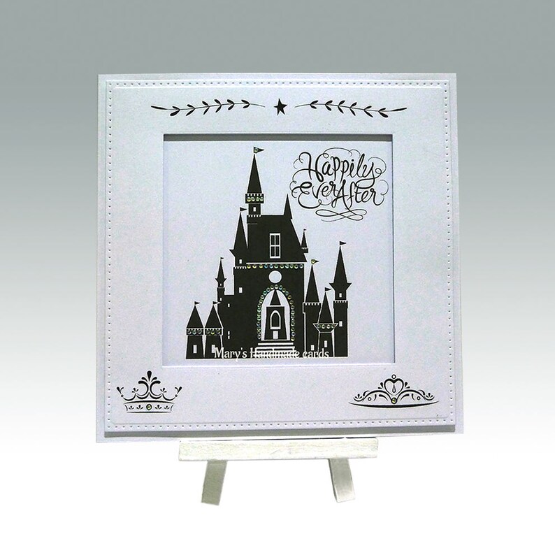 Pu&ograve; includere: Un biglietto bianco con una silhouette nera di un castello e il testo "Happily Ever After" in scrittura nera. Il biglietto &egrave; su un piccolo cavalletto bianco.