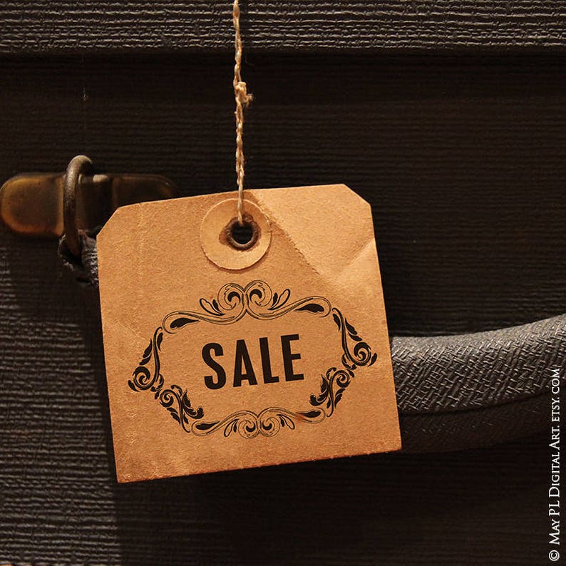 May include: A brown paper tag with the word "SALE" in a black decorative frame, hanging from a string. The tag is attached to a dark gray textured surface, possibly a suitcase or bag.