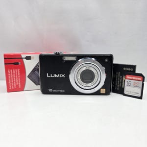 Op de afbeelding: Een zwarte Lumix 10 megapixel digitale camera met een zilveren Leica-lens, vergezeld van een zwarte Panasonic-accu, een 16GB Evo Plus-geheugenkaart en een rood-witte productdoos met tekst over het opladen van lithium-ionbatterijen.