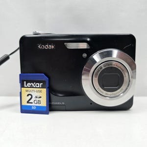 Könnte beinhalten: Eine schwarze Kodak Digitalkamera mit silbernem Objektivring, der die Aufschrift Kodak AF 3x Optical Aspheric Lens 32mm-96mm trägt. Eine blaue Lexar 2GB SD-Speicherkarte befindet sich vor dem Kameragehäuse. Die Kamera verfügt über einen Blitz und eine Handschlaufe.