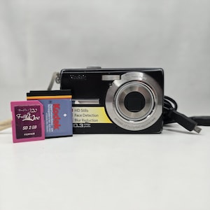Op de afbeelding: Een zwarte Kodak digitale camera met een zilveren lensring, een roze 2 GB Fujifilm SD-kaart en een blauwe en oranje Kodak oplaadbare batterij. De camerabehuizing bevat tekst zoals HD Stills, Face Detection, Blur Reduction en 10.3 mega pixels.