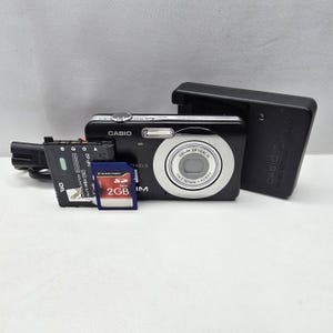 Könnte beinhalten: Eine schwarze Casio Exilim Digitalkamera mit silbernem Objektivring, begleitet von einem schwarzen Akkuladegerät, einem schwarzen NP-60 Akku und einer blauen Silicon Power 2GB SD-Speicherkarte. Die Kamera verfügt über einen Blitz.