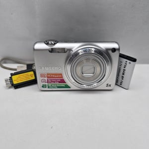 Op de afbeelding: Een zilveren Samsung digitale camera met een 5x zoomlens, 14,2 megapixels en HD-video-opname. De camera wordt vergezeld door een zwarte USB-oplaadkabel en een rechthoekige batterij met het label 3,7V 1200mAh 4,4Wh.
