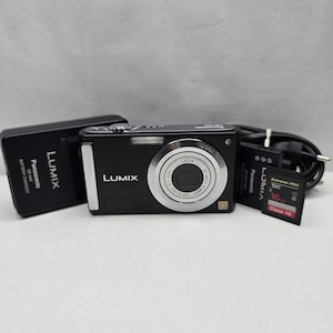 Op de afbeelding: Een zwarte Panasonic Lumix digitale camera met een Leica DC Vario-Elmarit lens, een Panasonic DE-A40 batterijlader, een Panasonic batterijpack, een zwarte stroomkabel en een SanDisk Extreme Pro 16GB Class 10 SD-geheugenkaart met rood label.
