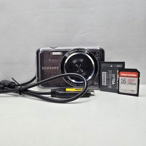 May include: A black Samsung digital camera with a 5x zoom lens, a black BP70A battery, a 16GB EVO Plus memory card, and a black USB charging cable. The camera body features a subtle glittery pattern and the Samsung logo on the front.
