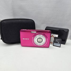 Puede incluir: Una cámara digital Sony Cyber-shot de color rosa brillante con un anillo de lente plateado, acompañada de un estuche negro con cremallera, un cargador de batería, una batería recargable y una tarjeta de memoria SD Sony de 2GB.