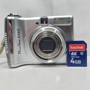 Op de afbeelding: Een zilverkleurige Canon PowerShot A560 digitale camera met een 4x optische zoomlens en een blauwe SanDisk 4GB SDHC-geheugenkaart. De camerabehuizing toont de modelnaam en de lens bevat de tekst 5.8-23.2mm 1:2.6-5.5.