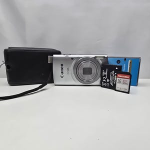 Könnte beinhalten: Eine silberne Canon IXUS Digitalkamera mit schwarzer Tragetasche, einem schwarzen wiederaufladbaren Akku und einer 16GB Professional EVO Plus Speicherkarte. Die Kamera verfügt über ein Frontobjektiv, einen Blitz und den Markennamen Canon IXUS.