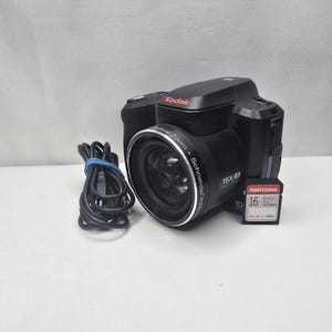 Könnte beinhalten: Eine schwarze Kodak Digitalkamera mit einem Schneider-Kreuznach Objektiv mit 15-fachem optischen Zoom, einem aufgewickelten schwarzen USB-Kabel und einer 16 GB EVO Plus Speicherkarte. Das Kameragehäuse zeigt den Markennamen Kodak in Rot.