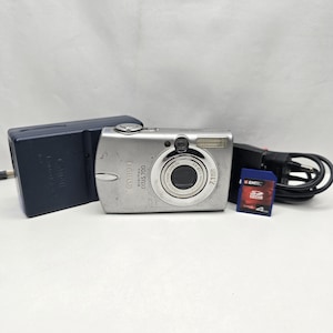 Könnte beinhalten: Eine silberne Canon Digital IXUS 700 Digitalkamera mit einem 7,1-Megapixel-Objektiv, begleitet von einem dunkelblauen Akkuladegerät, einem schwarzen Netzkabel und einer blauen Emtec 4GB SDHC-Speicherkarte. Das Kameragehäuse hat ein Metallfinish.