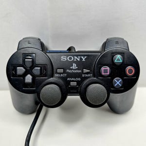Op de afbeelding: Een zwarte bedrade Sony PlayStation 2 DualShock 2 gamecontroller met een richtingspad, twee analoge sticks, vier actieknoppen met driehoek-, cirkel-, vierkant- en kruissymbolen, plus gelabelde Select-, Start- en Analog-knoppen.