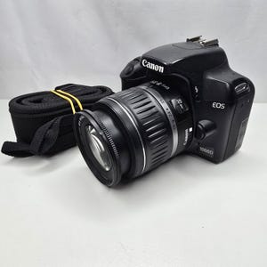Op de afbeelding: Een zwarte Canon EOS 1000D digitale spiegelreflexcamera met een bevestigde EFS 18-55mm lens. De camerabehuizing heeft het Canon-logo aan de bovenkant en de modelnaam 1000D aan de voorzijde. Een zwarte camerariem met een geel elastiek ligt ernaast.