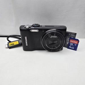 May include: A black Samsung WB690 digital camera featuring a Schneider-Kreuznach lens, a silver shutter button, and a mode dial. Included are a black battery labeled SLB-10A, a 4GB SanDisk SDHC memory card, and a black USB charging cable with a yellow label.