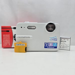 Peut inclure: Un appareil photo numérique Olympus blanc avec une étanchéité de 3m, un objectif et un flash. Il est accompagné d'un adaptateur microSD jaune Olympus MASD-1, d'une batterie lithium-ion argentée et d'une boîte d'emballage rouge et blanche.