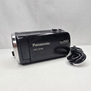 Può includere: Una videocamera Panasonic HDC-SD40 nera con bordo dell'obiettivo argentato e marchio Full HD 1920x1080 sul lato. Il dispositivo include un cavo di alimentazione nero avvolto posizionato a destra dell'unità principale, che mostra il nome del modello.